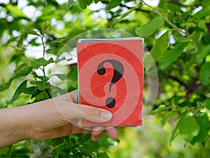 A woman holding a stack of colorful paper notes with question marks on them in her hand against a background of foliage