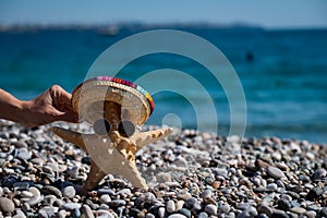 Woman holding a starfish in a sombrero and sunglasses on a pebble beach by the sea.