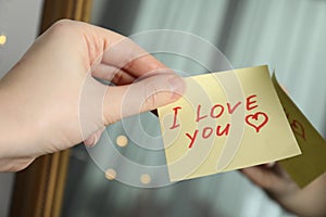 Woman holding sticky note with phrase I Love You near mirror, closeup