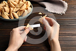 Woman holding tasty fortune cookie and paper with prediction at wooden table, closeup. Space for text