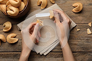 Woman holding tasty fortune cookie and paper with prediction at wooden table, top view. Space for text