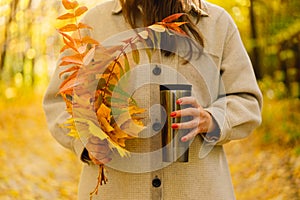 A woman is holding a thermo cup and a bouquet of autumn leaves. Autumn outdoor activity.