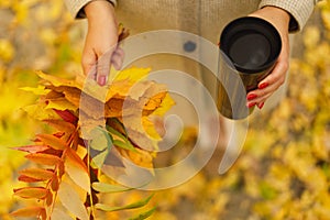 A woman is holding a thermo cup and a bouquet of autumn leaves. Autumn outdoor activity.