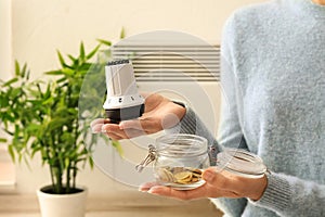 Woman holding thermostat and glass jar with coins at home. Heating saving concept