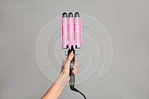 Woman holding triple curling hair iron on light grey background, closeup