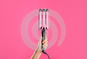 Woman holding triple curling hair iron on pink background, closeup