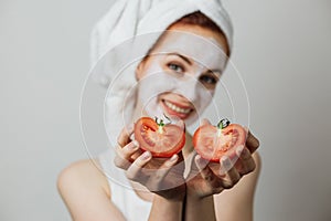 Woman holding two halves of tomato near her face with white clay mask on face