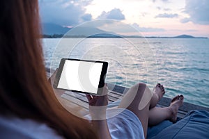 A woman holding and using black tablet with blank white desktop screen while sitting by the sea with blue sky background