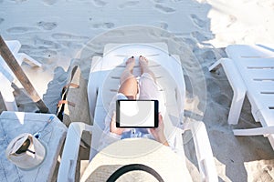 A woman holding and using a black tablet pc with blank desktop screen while laying down on beach chair on the beach