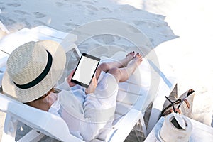 A woman holding and using a black tablet pc with blank desktop screen while laying down on beach chair on the beach