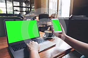 A woman holding and using mockup mobile phone with blank green screen and laptop on wooden table