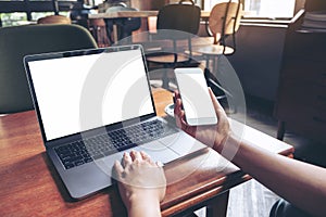 Woman holding and using mockup mobile phone with blank white screen and laptop on wooden table in cafe