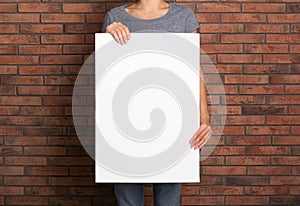 Woman holding white blank poster near red brick wall, closeup. Mockup for design