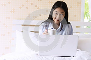 Woman holding white cup of hot coffee or tea while sitting on the bed in the morning. Teenager girl long hair