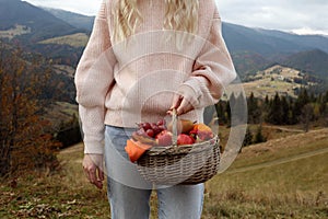 Woman holding wicker picnic basket with fruits and autumn leaves in mountains, closeup