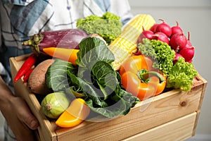 Woman holding wooden crate full of fresh vegetables on light background, closeup