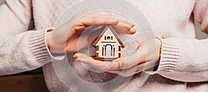 Woman holding a wooden house with her hands with the sun on a light pink background. Sweet home