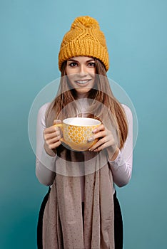 Woman holding yellow cup of tea wearing scarf and winter yellow hat, smiling at the camera