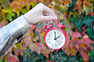 A woman holds an alarm clock against the background of autumn leaves. Reminder to switch to winter time