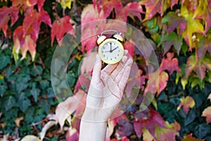 A woman holds an alarm clock against the background of autumn leaves. Reminder to switch to winter time