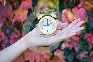 A woman holds an alarm clock against the background of autumn leaves. Reminder to switch to winter time