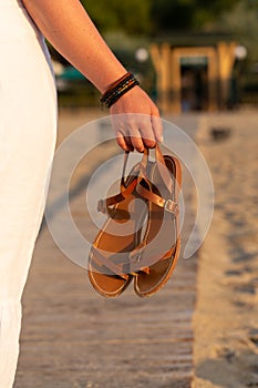 Woman holds brown leather sandals in her hand and looks at the beach hotel