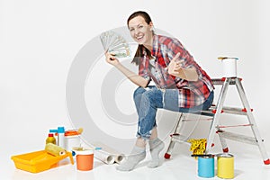 Woman holds bundle of dollars, cash money, sits on ladder with instruments for renovation apartment isolated on white