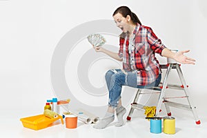 Woman holds bundle of dollars, cash money, sits on ladder with instruments for renovation apartment isolated on white