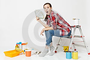 Woman holds bundle of dollars, cash money, sits on ladder with instruments for renovation apartment isolated on white