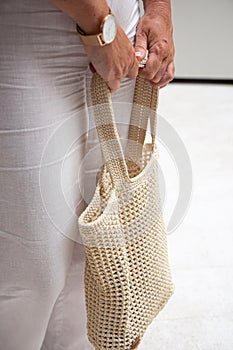 Woman holds a handmade beige knitted bag in her hand near her legs outdoors. Sustainable shopping. Wasteless lifestyle