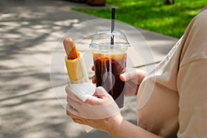 woman holds a hot dog and black ice coffee in her hands while walking in park. Street food