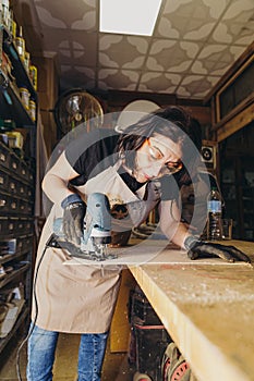 Woman holds jigging machine in her hand while cutting wooden plate in workshop photo