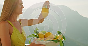 A woman holds a lemon up to the sunlight in sunlit setting. Girl examines and sniffing the fruit carefully, bowl with