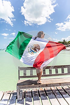 Woman holds Mexican flag behind her back as she stands on a bench on a pier next to the Carribean Sea