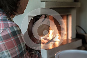 Woman with holds a mug of hot drink in her hands, looks at the flame in an open fireplace.