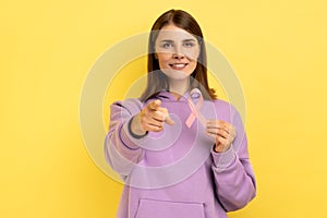 Woman holds pink ribbon near her chest supporting another women, cancer awareness pointing to camera