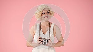 A woman holds up an old vintage camera. Woman in the image of Marilyn Monroe in studio on pink background close up.