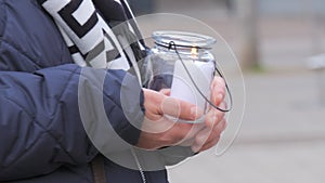 A woman holds a white large church candle in a glass candle holder outside in the cold season
