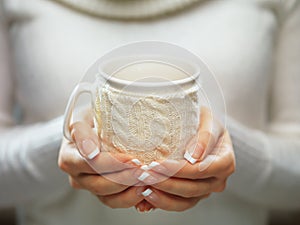 Woman holds a winter cup close up. Woman hands with elegant french manicure nails design holding a cozy knitted mug.