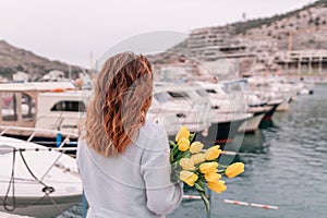 Woman holds yellow tulips in harbor with boats docked in the background., overcast day, yellow sweater, mountains