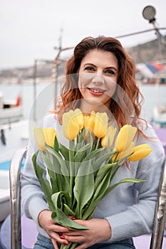 Woman holds yellow tulips in harbor with boats docked in the background., overcast day, yellow sweater, mountains