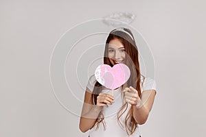 Woman with holy nimbus holding pink paper heart on stick and pointing to camera, demonstrating love.