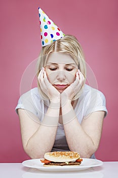 A woman in home clothes and a festive cap sits sad in front of a hamburger on a plate. Lonely birthday in isolation period on the