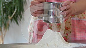 A woman in a home kitchen sifts flour through a metal sieve. Sifted flour falls on the countertop with a cute kitchen