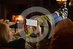 Woman At Home Lying On Sofa In Lounge With Cosy Fire Streaming Onto Blank Screen Mobile Phone