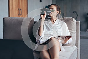 Woman at home in a nightgown with a magazine, a glass and patches on her face.