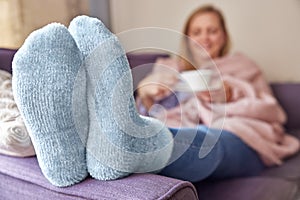 Woman At Home Wearing Cosy Warm Socks And Wrapped In Blanket Lying On Sofa Drinking Cup Of Tea