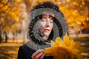 Woman in a hood with fur in the autumn forest. A girl in warm outerwear holds a bouquet of maple leaves