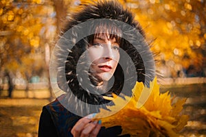 Woman in a hood with fur in the autumn forest. A girl in warm outerwear holds a bouquet of maple leaves