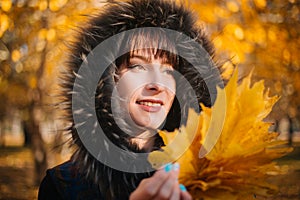 Woman in a hood with fur in the autumn forest. A girl in warm outerwear holds a bouquet of maple leaves